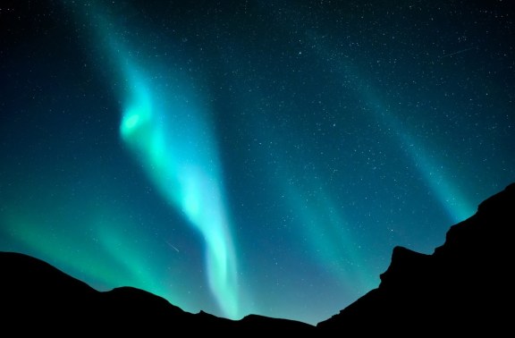 The image showcases the stunning Aurora Australis, commonly known as the Southern Lights, casting a brilliant display of green and blue lights across a star-studded night sky. The silhouette of a rugged, dark mountainous landscape adds dramatic contrast to the luminous celestial phenomenon. This natural light display occurs in the high-latitude regions around the Antarctic and is caused by the interaction of the Earth’s magnetic field with charged particles from the sun. The scene captures the ethereal beauty of one of nature's most spectacular shows, visible in the polar regions of the Southern Hemisphere.