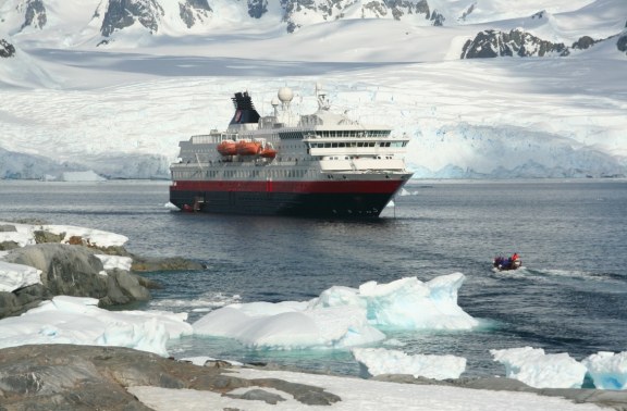 A large red and black cruise ship anchored near the Antarctic coast, with passengers in a small zodiac boat nearby, surrounded by icy blue waters and snow-covered landscapes