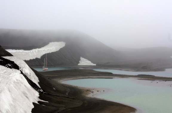 A small red sailboat anchored at Deception Island, with dramatic black and white snowy hills rising in the background.