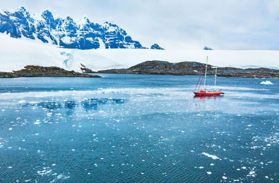 A red sailboat anchored in a calm bay with icy waters scattered with small ice floes, set against a backdrop of snow-covered mountains in the Antarctic Peninsula.